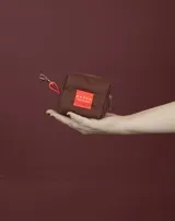 Monedero pequeño de tela color beige con cierre azul y tira de agarre color verde. Incluye un parche rectangular rojo con logo y gancho metálico para colgar.