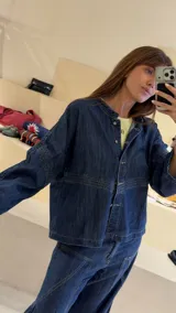 Camisa de denim azul con cuello mao, diseño holgado, mangas largas y cierre frontal con botones metálicos. Cuenta con costuras decorativas pespunteadas en el delantero.