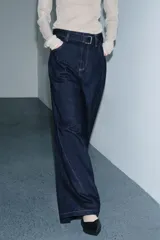 Jeans de tiro alto con pierna ancha, confeccionados en denim azul oscuro. Presentan cinco bolsillos, trabillas en la cintura y un cinturón ajustable del mismo tejido con hebilla metálica. Incluyen pespuntes marcados a tono y cierre frontal con cremallera y botón.