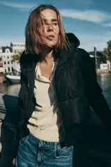 Chaqueta acolchada negra con capucha ajustable, cuerpo con aislamiento térmico y mangas largas en tejido técnico efecto neopreno. Presenta cierre frontal con cremallera, bolsillos laterales y bajo ajustable con cordones elásticos.