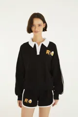 Buzo negro de felpa con cuello polo y solapa blanca, mangas largas con franjas blancas en los laterales y estampado en el pecho con la palabra "RIO" y detalles gráficos.
