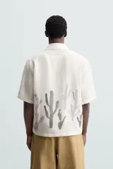 Camisa de calce relajado confeccionada en tejido ligero de lino y viscosa, con cuello solapa, manga corta y cierre frontal de botones. Presenta un diseño estampado de cactus en color gris en la parte inferior sobre un fondo blanco roto.
