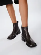 Botas tipo chelsea de cuero color marrón oscuro con acabado brillante. Presentan punta cuadrada con detalle de puntera metálica, elásticos laterales y taco cuadrado bajo.