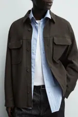 Sobrecamisa relaxed fit color beige con tejido elástico, cuello solapa, manga larga con puños abotonados, bolsillos de plastrón con solapa en el pecho, bolsillos laterales de vivo en la cadera y cierre frontal de botones.