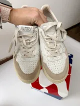 Championes Autry de diseño retro, color blanco con detalles en gamuza beige y suela de tono tostado, con cordones claros y logo lateral de la marca con la bandera de Estados Unidos.