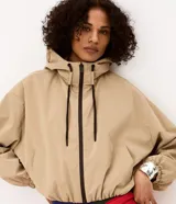 Campera cortavientos de poliéster color beige, modelo corto con capucha ajustable mediante cordón oscuro, cierre frontal por cremallera oscura, bolsillos laterales y puños y ruedo elásticos.