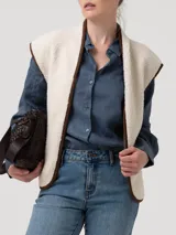 Chaleco de polar tipo corderito en color beige, con cierre frontal y detalles de ribetes en color marrón. Presenta bolsillos laterales y un diseño sin mangas.