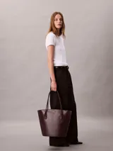 Cartera tote Calvin Klein de color marrón oscuro, con doble asa de hombro, cierre de cremallera superior y monograma metálico dorado en la parte frontal.