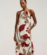 Vestido midi de corte evasé, con escote halter y abertura frontal en el busto, anudado en la parte superior. Presenta un estampado grande de flores y hojas en tonos rojo oscuro, beige y negro sobre fondo crudo. El diseño incluye un volante fruncido en el ruedo inferior.