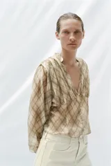 Camisa de manga larga con escote en V y cierre frontal con botones. Presenta un estampado de cuadros en tonos beige y marrón, con detalles de pliegues en el delantero y puños ajustados.