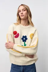 Sweater de punto color crudo con diseño de flores y formas geométricas bordadas en colores vibrantes. Presenta cuello redondo, mangas largas con detalles de líneas laterales en contraste y corte holgado.