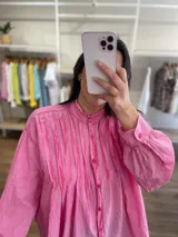 Camisa de algodón color amarillo con diseño de línea italiana, corte holgado y asimétrico. Presenta cuello camisero, cierre frontal con botones y detalle de pliegues verticales en el pecho. Cuenta con mangas largas y puños con botón.