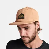 Gorra color beige con visera plana y parche con el logo de la marca Roark bordado en el frente.