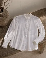 Camisa de manga larga color blanco, con cuello alto y detalle de volados en el cuello y a lo largo de la botonadura frontal. Presenta puños con volados y cierre de botones.