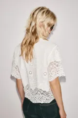 Camisa blanca de manga corta y cuello solapa, con detalles de bordados perforados y encaje en el borde inferior y las mangas. Cuenta con cierre frontal de botones.