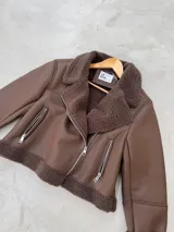 Campera tipo gamulán de cuero sintético en color marrón chocolate, con interior y solapas de corderito sintético al tono. Presenta cierre frontal cruzado y bolsillos laterales con cierre metálico.