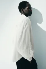 Blusa oversize blanca de algodón y viscosa, con cuello camisero, tapeta de botones ocultos y detalles fruncidos en el canesú delantero y trasero. Presenta mangas globo largas con puños abotonados.