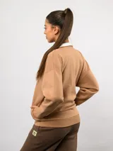 Buzo de felpa color tostado con cuello alto y medio cierre frontal. Presenta un diseño de corte holgado con hombros caídos, puños y ruedo acanalados, y el logo de la marca bordado en el pecho.