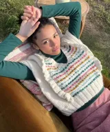 Chaleco de punto blanco color marfil con escote en V, tejido a mano en lana merino. Presenta detalles de pequeños vuelos en las sisas y franjas horizontales con bordados texturizados de colores vibrantes en el frente.