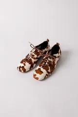 Zapato tipo flat de estilo acordonado, confeccionado en cuero con pelo con estampado de manchas tipo vaca en tonos marrón y blanco.