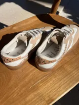 Zapatillas urbanas de cuero color beige y blanco, con tachas plateadas en los laterales y suela de goma color caramelo. Diseñadas en Uruguay y producidas en Brasil.