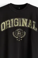 Remera negra de punto pesado de algodón con cuello redondo, hombros caídos y diseño holgado. Presenta un estampado frontal estilo universitario con la palabra "ORIGINAL" en tono desgastado y un gráfico circular rodeado de laureles.