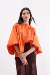 Camisa holgada de popelina de algodón con rayas verticales en tonos rosado, gris y blanco. Presenta cuello alto con volado, hombros caídos, mangas anchas abullonadas y cierre frontal con botones.