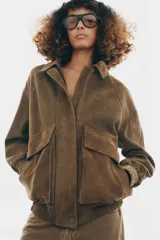 Chaqueta oversize confeccionada en piel de ante, con cuello solapa, manga larga con puños elásticos y bajo elástico. Presenta bolsillos de plastrón con solapa en el frente y cierre frontal con cremallera y botones ocultos.