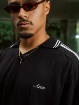 Chomba de tejido de punto cerrado color blanco, con cuello polo y mangas cortas. Presenta un bordado sutil en el frente y tres franjas negras en los hombros.