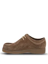 Zapato tipo wallabee de Hush Puppies, confeccionado en cuero gamuzado color marrón. Presenta costuras a la vista en tono claro, cordones al tono y suela de goma texturizada. Incluye etiqueta lateral con la marca y bandera británica.