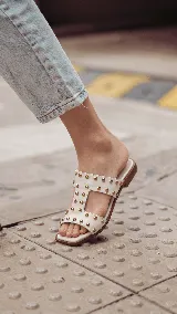 Sandalia baja estilo chinela de cuero color blanco off white, con tiras anchas decoradas con tachas metálicas doradas.