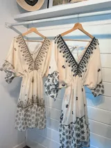 Vestido corto color crudo con bordado floral negro, escote en V, mangas 3/4 acampanadas y cintura ajustable con cordón.