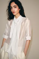 Camisa blanca de tejido semitransparente ZARA Woman Collection, con cuello redondo, manga corta y detalle de pañuelo integrado en el mismo tejido al cuello. Cierre frontal con botones.