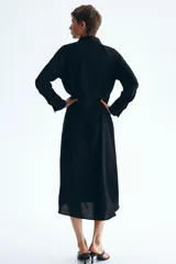 Vestido midi negro con cuello camisero y tapeta con botones ocultos, de mangas largas raglán con puños anchos y costura fruncida en la cadera con falda de vuelo leve.