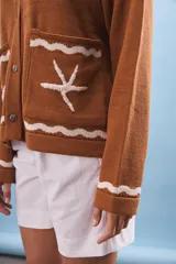 Cárdigan marrón de tejido de punto con cuello redondo y cierre frontal con botones. Tiene bolsillos frontales con diseño de olas y estrellas de mar en color blanco.