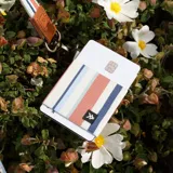 Billetera elástica Thread con diseño a rayas verticales en tonos azul, naranja y blanco. Incluye llavero metálico y logo de la marca en etiqueta tejida.