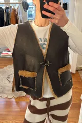 Chaleco reversible confeccionado en cuero genuino. Presenta un lado de piel de oveja gris con bolsillos de cuero negro y un lado de cuero liso gris. Diseño sin mangas con cierre frontal.