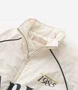 Campera bomber de microfibra color beige con estampado frontal de lettering 'PARIS 1985'. Presenta cierre frontal con cremallera, cuello alto, bolsillos funcionales y puños y ruedo ajustados con elástico.