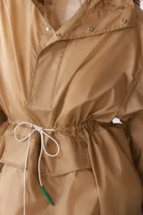Cazadora tipo anorak color beige, con capucha ajustable, manga larga y bolsillo frontal de plastrón con solapa. Cuenta con cordones elásticos ajustables en la cintura y el bajo, y cierre frontal con botones a presión.