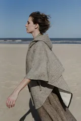 Capa amplia con capucha, confeccionada en lana fina sin teñir. Presenta cierre frontal, bolsillos delanteros y botones laterales que permiten ajustar la forma de las mangas.