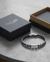 Pulsera masculina con diseño de eslabón central de acero inoxidable con patrón geométrico griego, flanqueado por secciones de acero y correa de cuero gris.