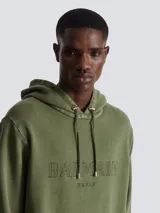 Hoodie de algodón color verde oliva con efecto vintage, presenta el logo de Balmain Paris bordado en el pecho, capucha con cordones ajustables y bolsillo tipo canguro frontal.