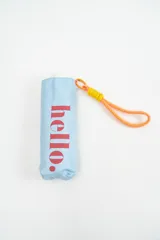 Paraguas plegable de color celeste con la palabra 'hello.' estampada en color rosado. Incluye una correa de mano naranja con detalle de hilo amarillo.