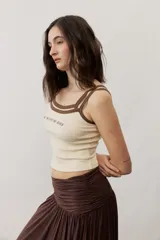 Musculosa de tejido rib color beige con detalles en marrón oscuro en el escote y doble tirante fino. Presenta un pequeño texto estampado en el pecho.