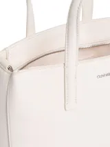 Cartera tipo tote pequeña de color blanco, con asas de mano fijas y correa ajustable opcional. Presenta un logo metálico de Calvin Klein en la parte delantera y un logo bordado en la correa de tela.