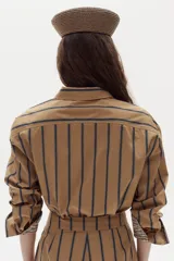Camisa boxy de popelina de algodón color tostado con diseño de rayas verticales oscuras, cuello camisero, botones frontales y mangas largas con puños estampados a cuadros.