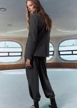 Pantalón de traje gris con sutil diseño de cuadros, tiro medio y pernera recta. Cuenta con trabillas para cinturón, pinzas delanteras, bolsillos laterales y cierre frontal con cremallera, botón y ganchos ocultos.