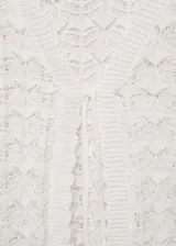 Cardigan de crochet color blanco, con cuello en V, manga larga y cierre frontal mediante lazos con borlas. Presenta un diseño calado con textura.