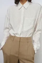 Camisa fluida de corte oversize confeccionada en mezcla de viscosa, con diseño de rayas verticales finas en tono beige sobre fondo blanco. Presenta cuello solapa, cierre frontal con botones, manga larga con puños y un bolsillo de plastrón en el pecho.