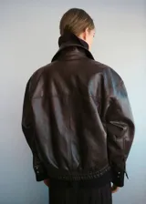 Chaqueta estilo bomber de cuero sintético en color marrón oscuro, con cuello camisero, cierre frontal de cremallera y botones a presión, bolsillos de parche con solapa y bajo elástico.
